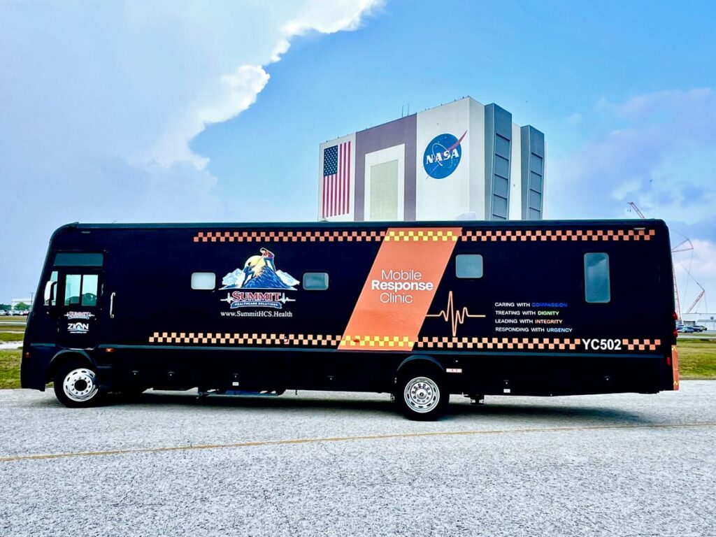 A mobile response clinic vehicle parked in front of a NASA building with an American flag, under a partly cloudy sky.