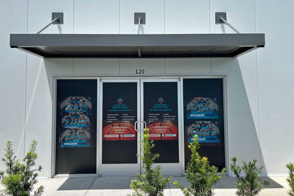 Double glass doors with dark coverings and promotional signs, under a metal awning on a white building; small shrubs line the sidewalk in front.