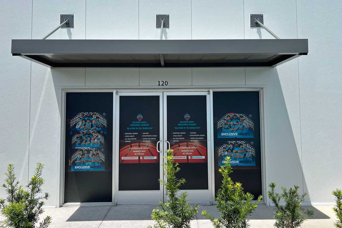 Double glass doors with dark coverings and promotional signs, under a metal awning on a white building; small shrubs line the sidewalk in front.