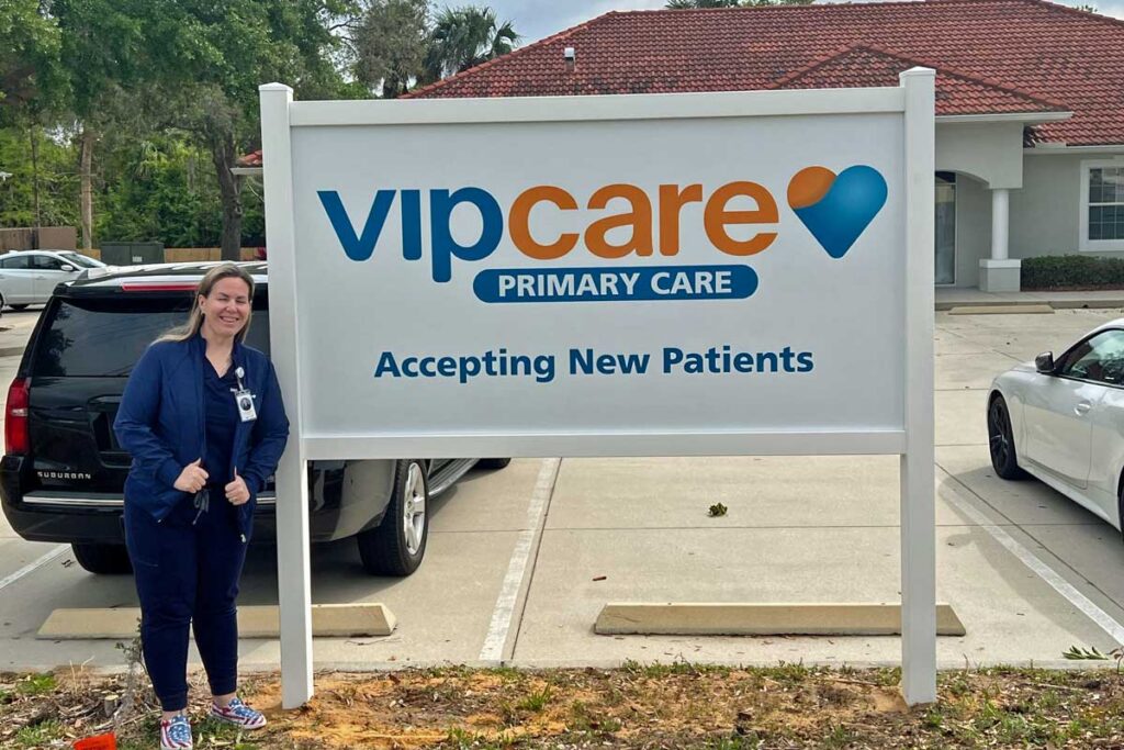 A person stands smiling next to a large sign that reads "VIPcare Primary Care - Accepting New Patients" in a parking lot.