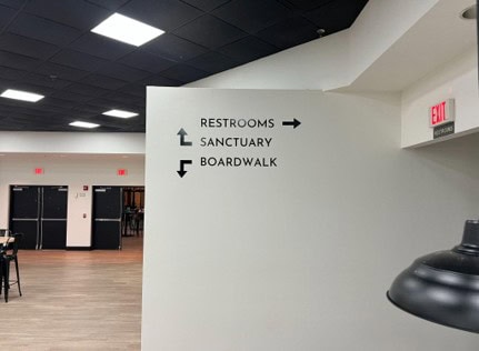 White wall with black directional signs indicating restrooms to the right, and sanctuary and boardwalk to the left.