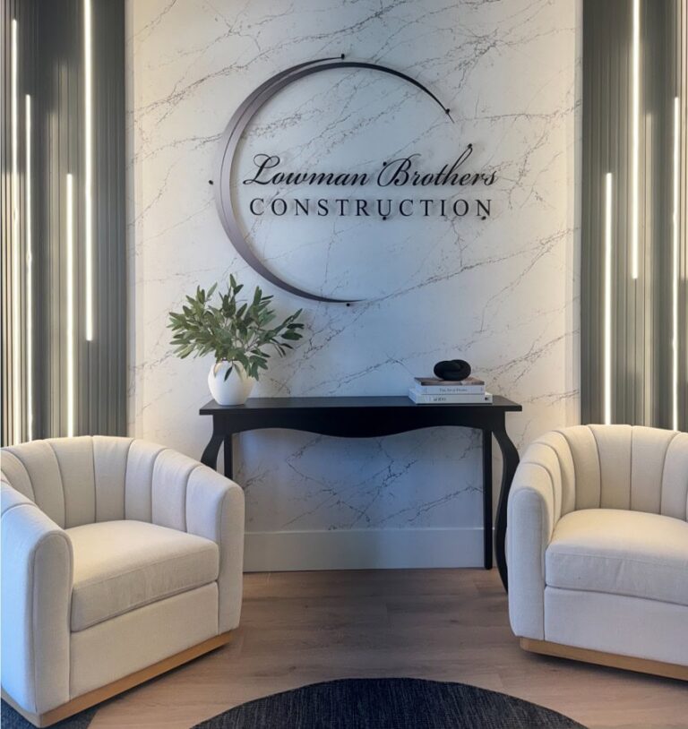 Two cream armchairs face a black console table with a plant, books, and décor. “Lawman Brothers Construction” sign is mounted on a marble-patterned wall in the background.