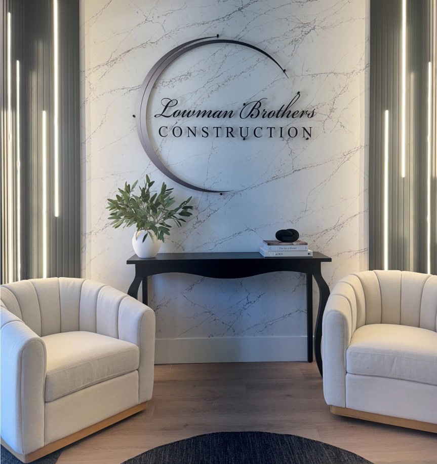 Two cream armchairs face a black console table with a plant, books, and décor. “Lawman Brothers Construction” sign is mounted on a marble-patterned wall in the background.