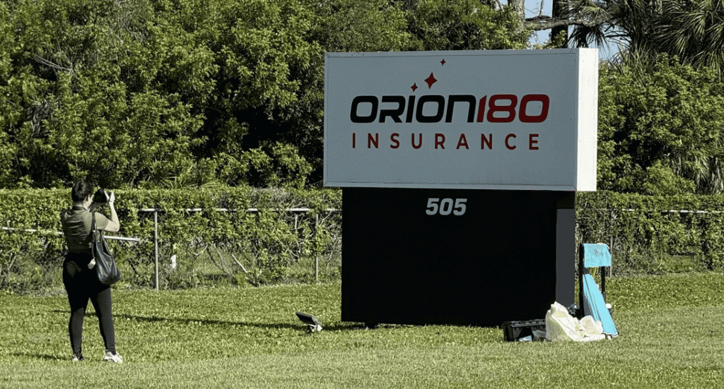 A person takes a photo of a large Orion180 Insurance sign on a grassy lawn, with trees and fencing in the background.