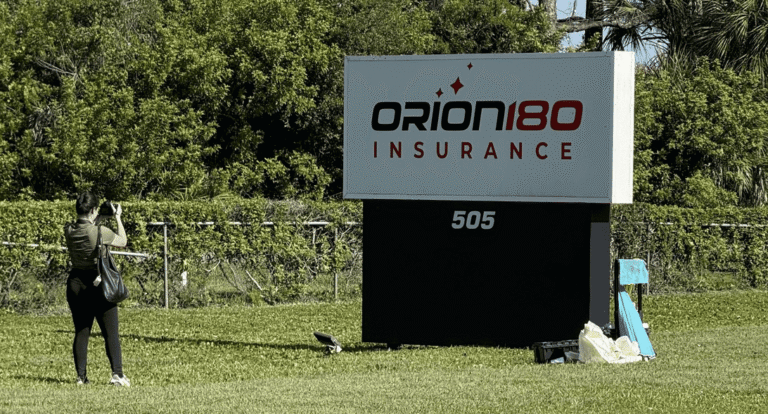 A person takes a photo of a large Orion180 Insurance sign on a grassy lawn, with trees and fencing in the background.
