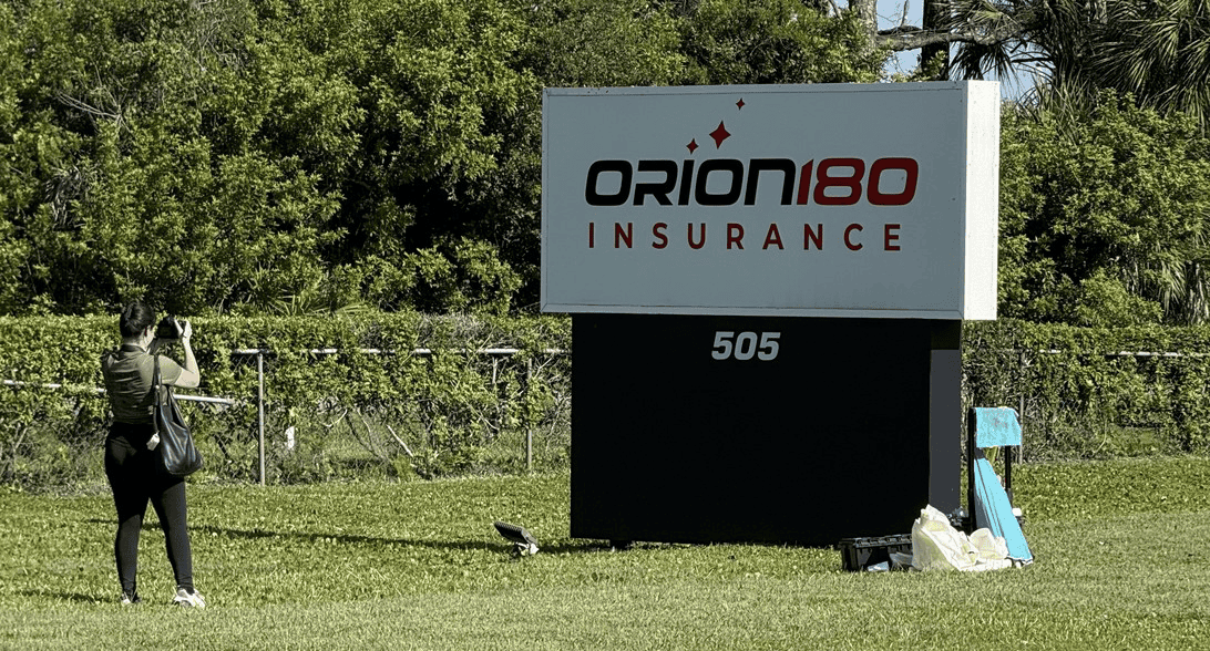 A person takes a photo of a large Orion180 Insurance sign on a grassy lawn, with trees and fencing in the background.