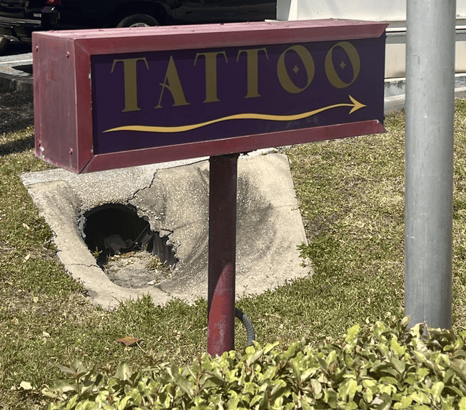 A maroon sign with yellow text reading “TATTOO” and an arrow points toward a sidewalk with a large, broken hole in the concrete.