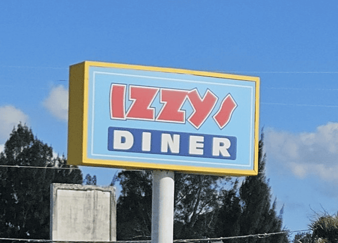 A rectangular sign reading “Izzy’s Diner” in bold red and white letters against a blue background stands outdoors with trees and a clear sky behind it.