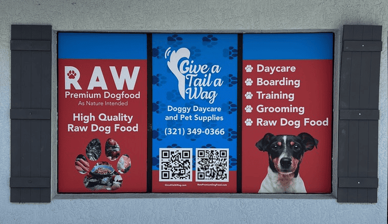 Storefront window display for Give a Tail a Wag featuring services like daycare, boarding, training, grooming, and raw dog food, with a QR code and photo of a black and white dog.