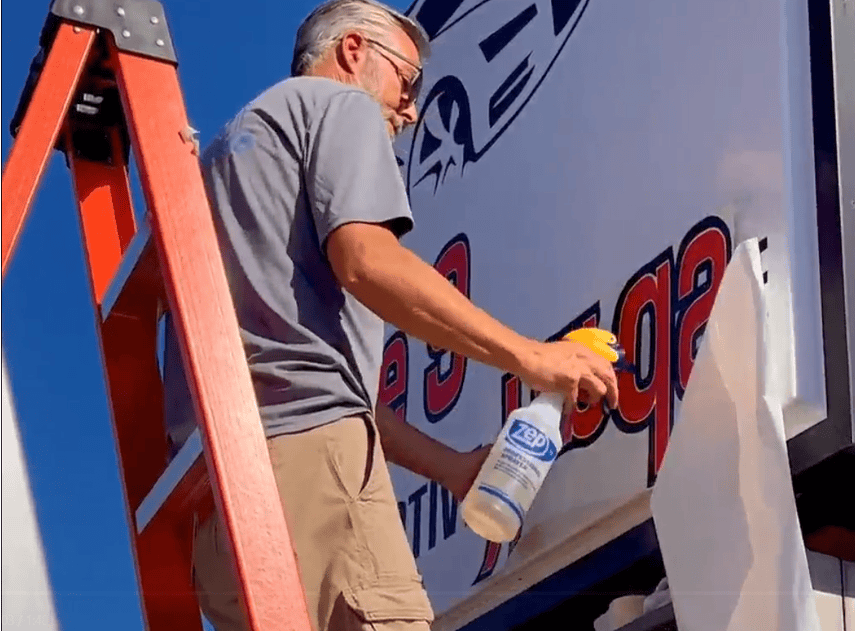A man on a ladder uses a spray bottle to clean or prepare the surface of a large outdoor sign with bold red and black text.