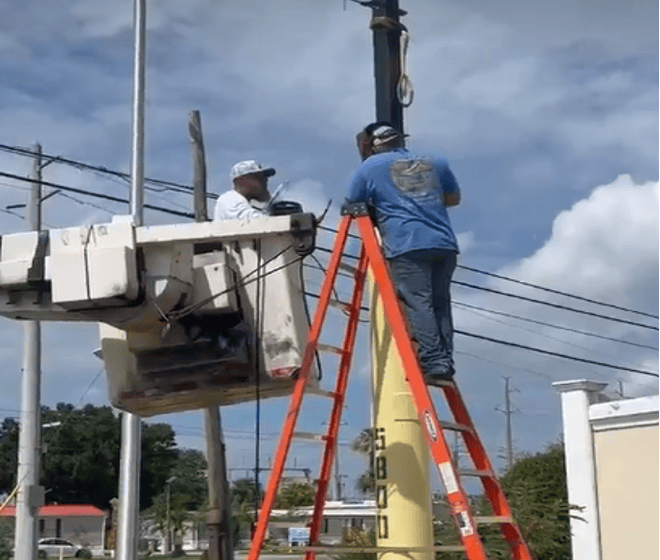 Two workers fix a streetlight; one stands on a red ladder, the other is in a bucket lift. Power lines and buildings are visible in the background under a partly cloudy sky.