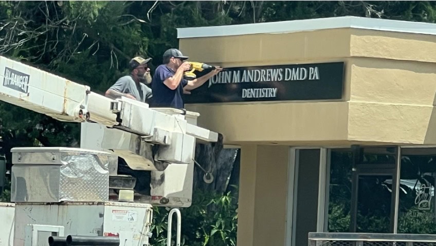 Two workers in a bucket lift install or remove a sign reading “JOHN M. ANDREWS DMD PA DENTISTRY” on the exterior of a building.