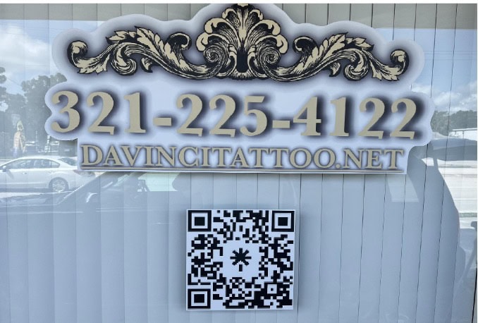 Window sign with ornate design, phone number 321-225-4122, website DAVINCITATTOO.NET, and a QR code; reflection of cars and trees in the background.