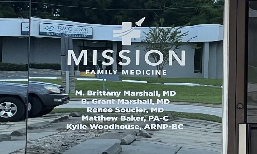 Glass door with “Mission Family Medicine” logo and the names of medical staff, reflecting a parked car and buildings across the street.