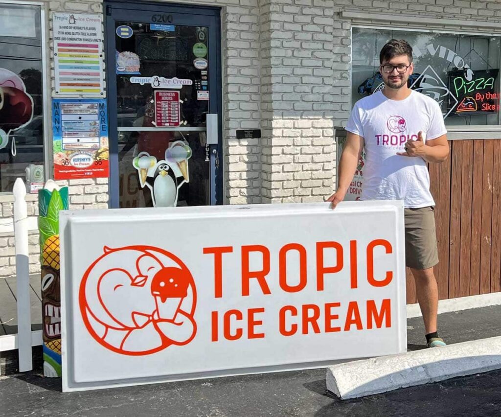 A man stands outside a storefront holding a large “Tropic Ice Cream” sign with a cartoon elephant logo.