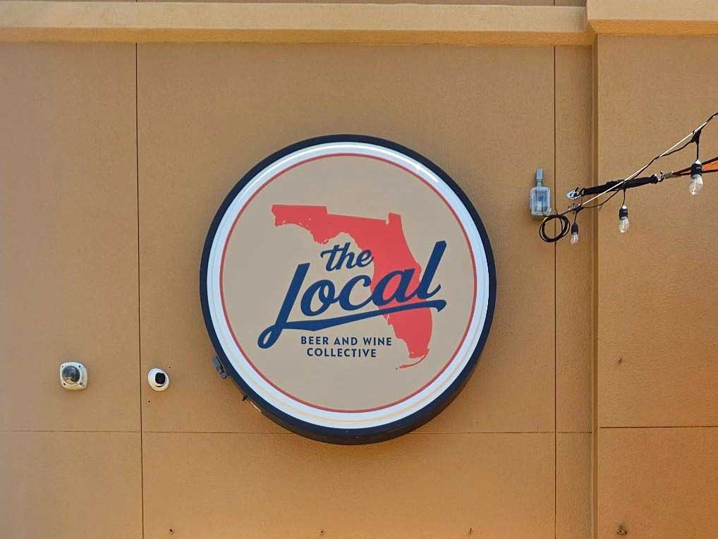 A round sign on a tan wall displays “The Local Beer and Wine Collective” with an outline of Florida in red and string lights hanging nearby.