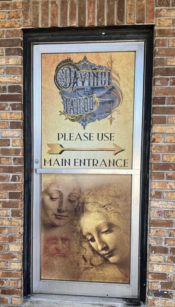 A door with “Da Vinci Tattoo” signage, artwork of two faces, and a sign that reads “Please use main entrance” with arrows pointing right, set in a brick wall.