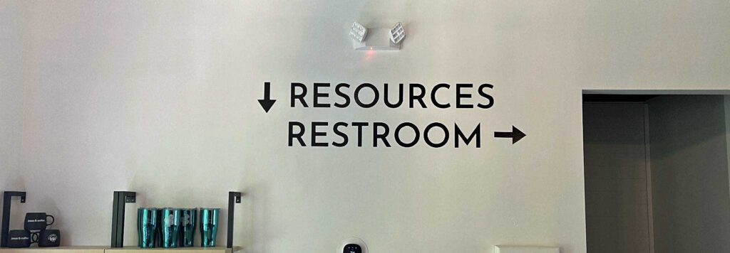 Black text on a wall points down for resources and right for restroom; shelves with teal containers are on the left.