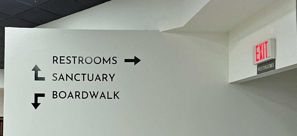 White wall with black directional signs pointing to restrooms (right), sanctuary (up left), and boardwalk (down left); an exit sign is above a hallway.