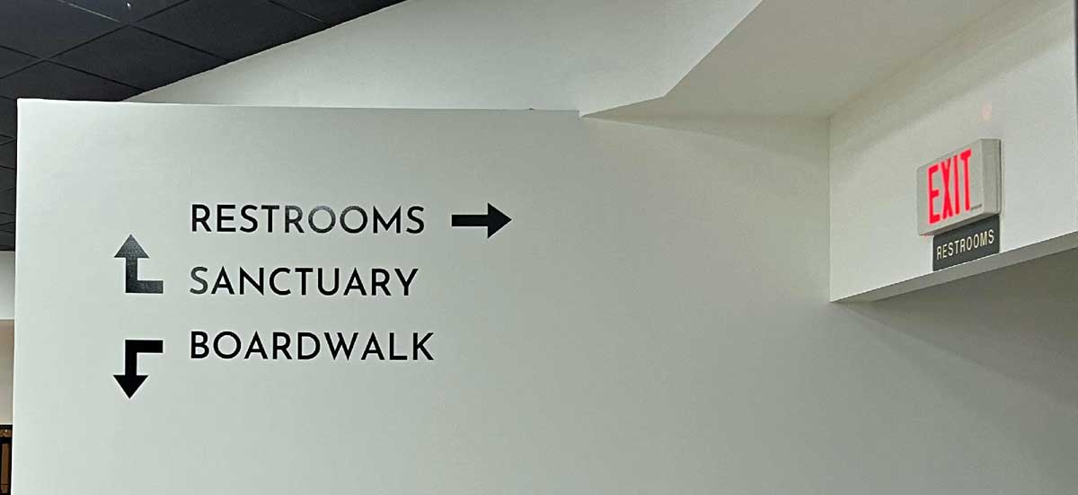 White wall with black directional signs pointing to restrooms (right), sanctuary (up left), and boardwalk (down left); an exit sign is above a hallway.